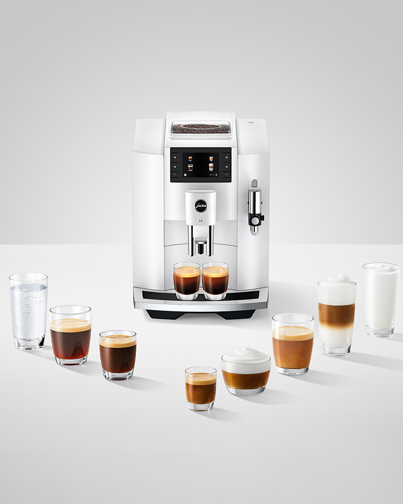 Abbildung: Weiße JURA E8 Kaffeemaschine mit verschiedenen zubereiteten Kaffee- und Milchspezialitäten auf einem weißen Tisch.