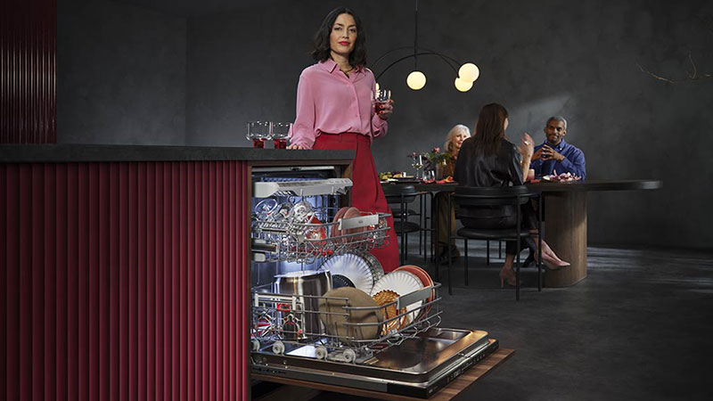 Abbildung: Frau mit Weinglas steht vor einem geöffneten AEG-Geschirrspüler voller gespülter Teller. Im Hintergrund sitzen Gäste an einem modern gedeckten Esstisch.