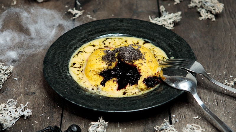 Abbildung: Herbstlicher Kürbis-Germknödel auf Vanillesoße, gefüllt mit dunkler Beerenfrucht, angerichtet mit Mohn und flüssiger Butter – serviert auf schwarzem Teller mit herbstlicher Deko.