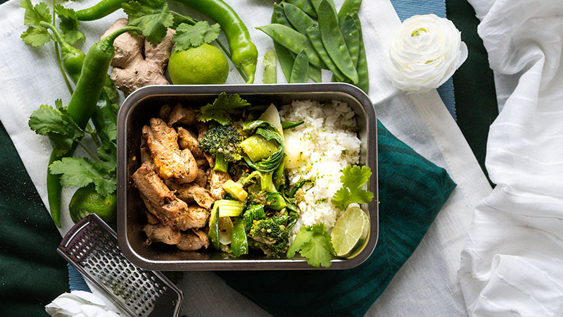 Abbildung: Draufsicht auf Edelstahl-Lunchbox mit Gemüse, Reis und Hähnchenfleisch, umgeben von frischen Zutaten wie Limetten, Ingwer, Koriander und Zuckerschoten auf gedecktem Tisch.