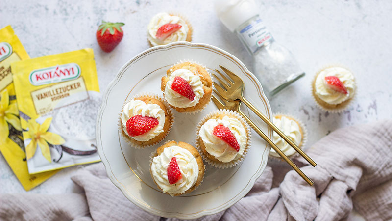 Abbildung: Fünf Erdbeer-Cupcakes auf einem weißen Porzellanteller mit goldenen Gabeln, daneben Vanillezucker von Kotányi, eine Prise Meersalz und lose liegende Erdbeeren