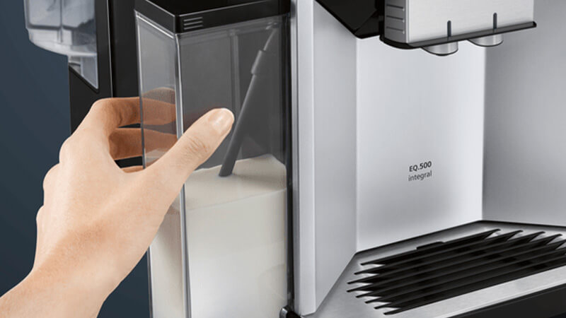 Abbildung: Hand greift seitlich an den transparenten Milchbehälter eines Siemens EQ.500 Kaffeevollautomaten – bequeme Entnahme für Reinigung oder Befüllen.